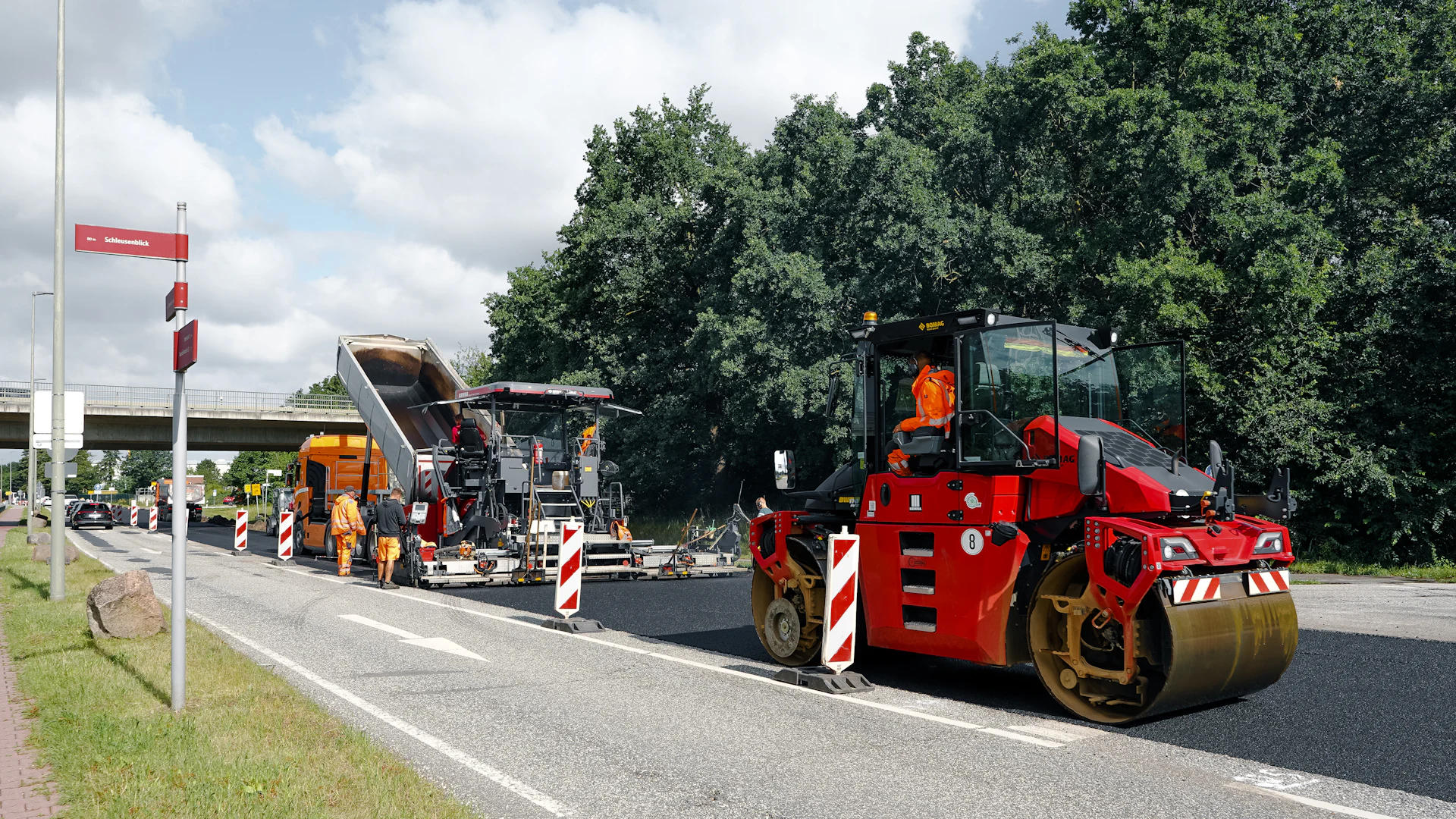 A red road roller and asphalt paving crew working on a road in daylight